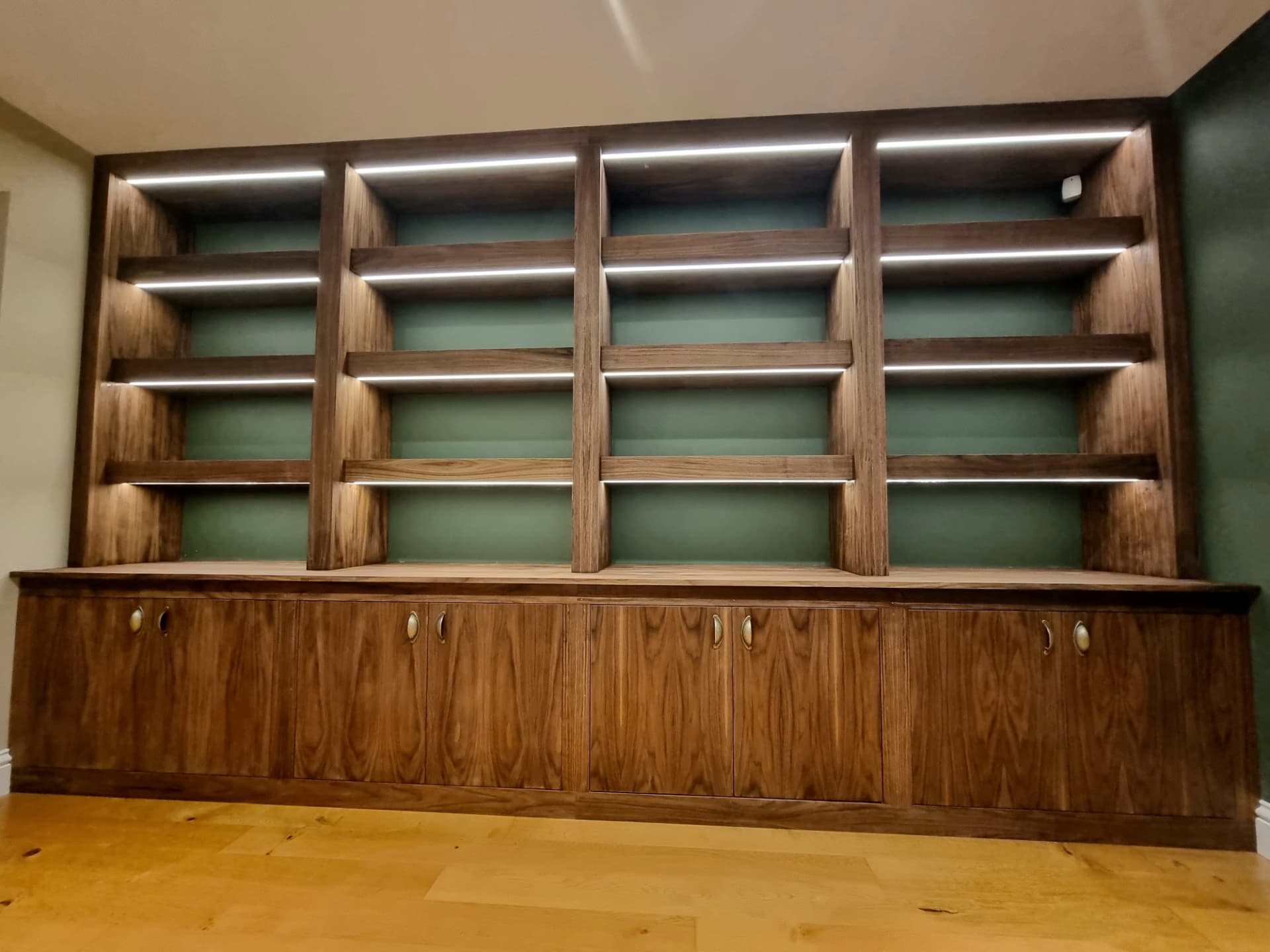 Fitted wall unit in dark walnut veneer with lower cupboards, oval brass drop handles and open lit shelving against a sage-green wall
