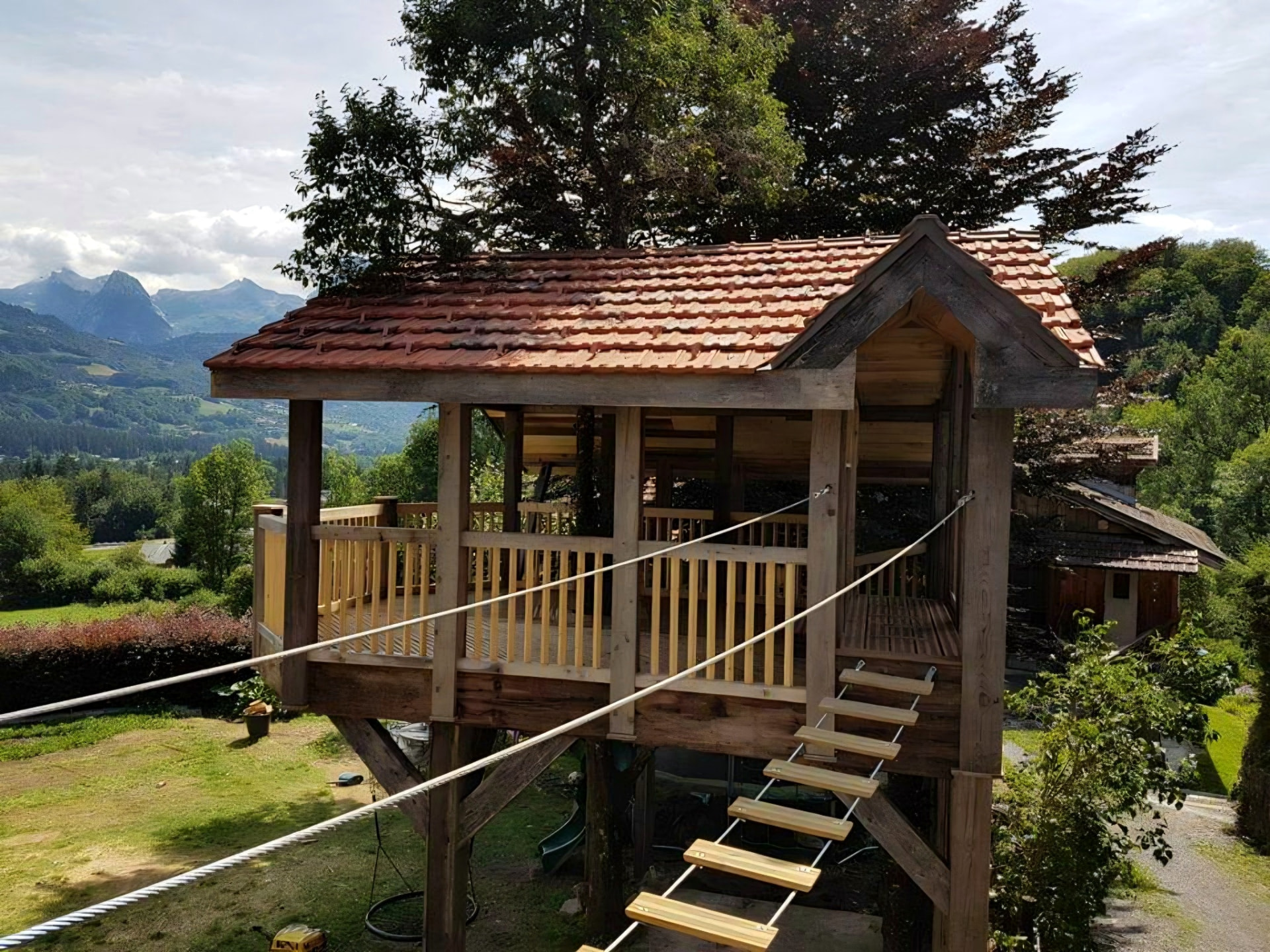 Timber treehouse with a red tiled roof and rope bridge, overlooking an alpine valley and mountains
