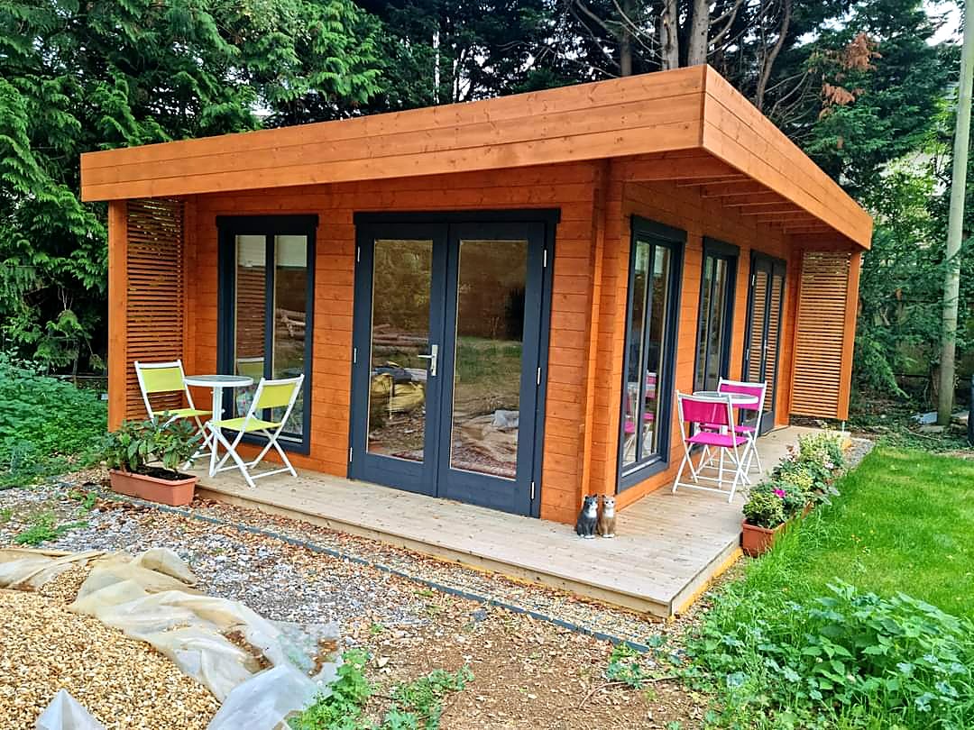 Timber garden room with a wide overhanging roof, dark grey French doors and tall windows, standing on a light timber deck