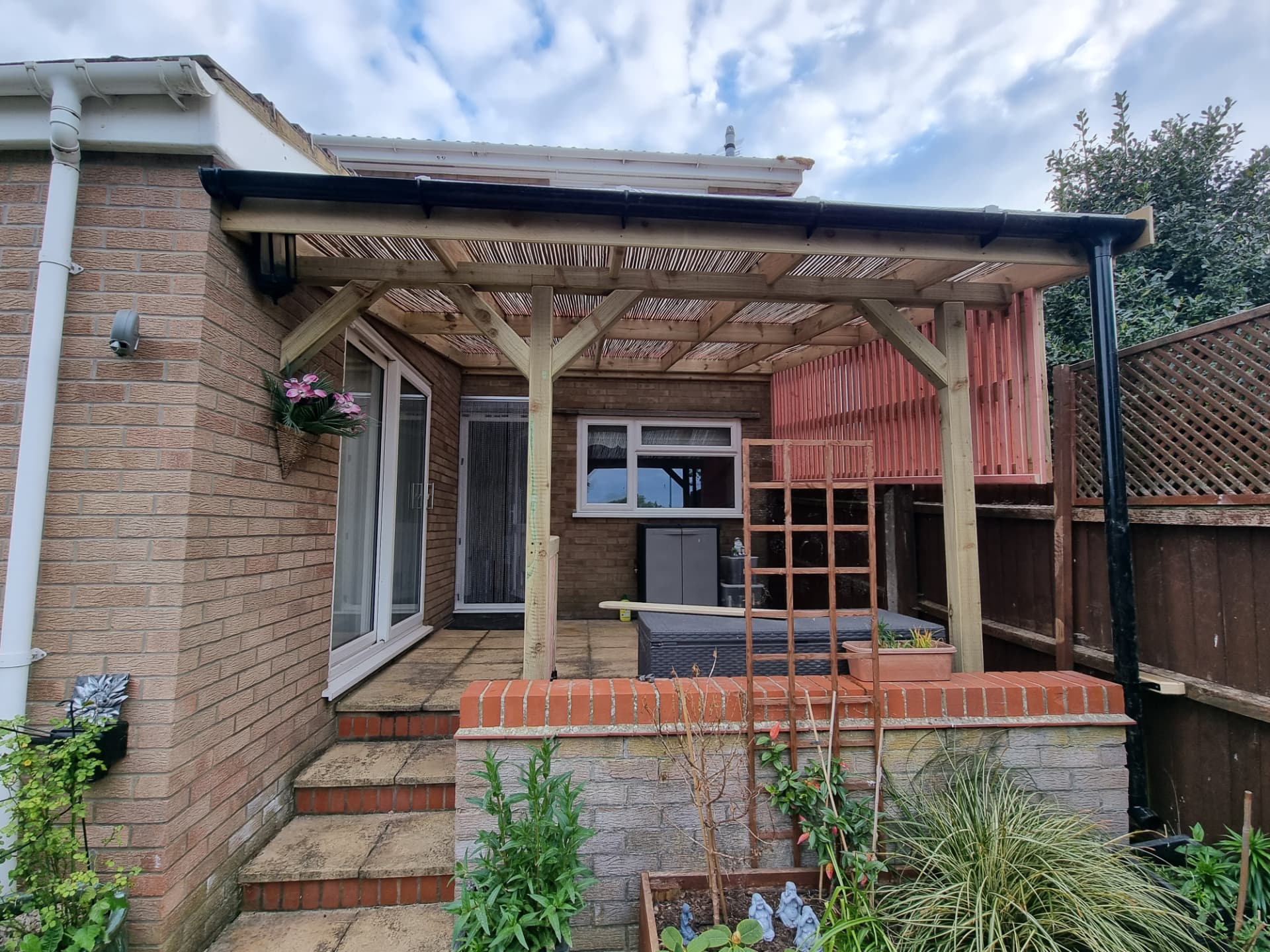 Lean-to timber pergola fixed to a brick house over a paved terrace, with a sloping reed-screened roof and black guttering