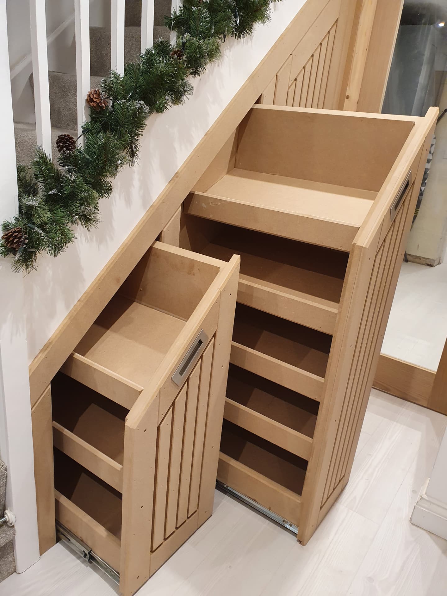 Two pull-out under-stairs shoe cabinets with stepped tops and recessed metal handles, under a staircase with white balusters