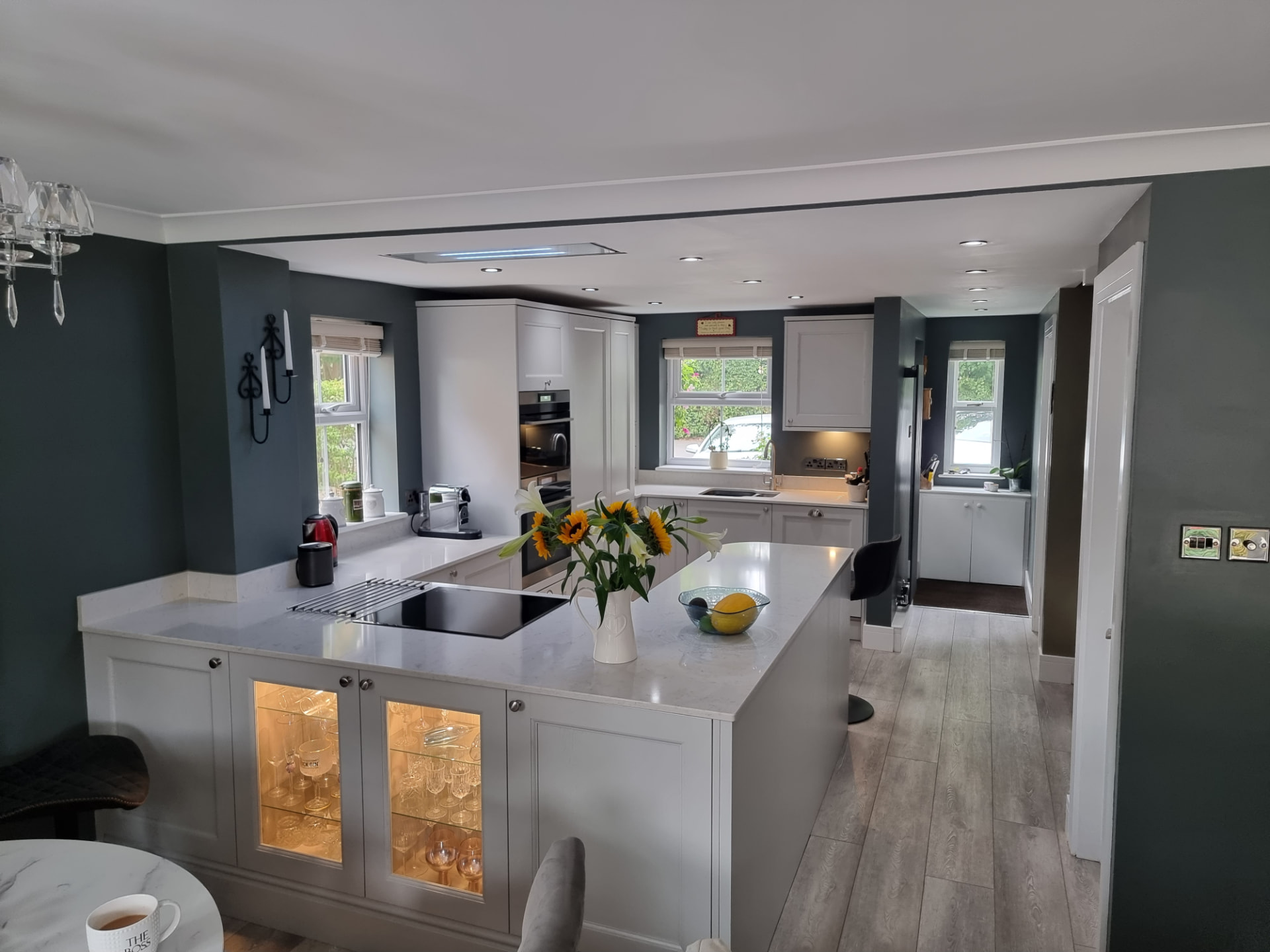 Dove-grey shaker kitchen with a quartz peninsula, lit glass-front display cabinets, an induction hob and a double oven tower