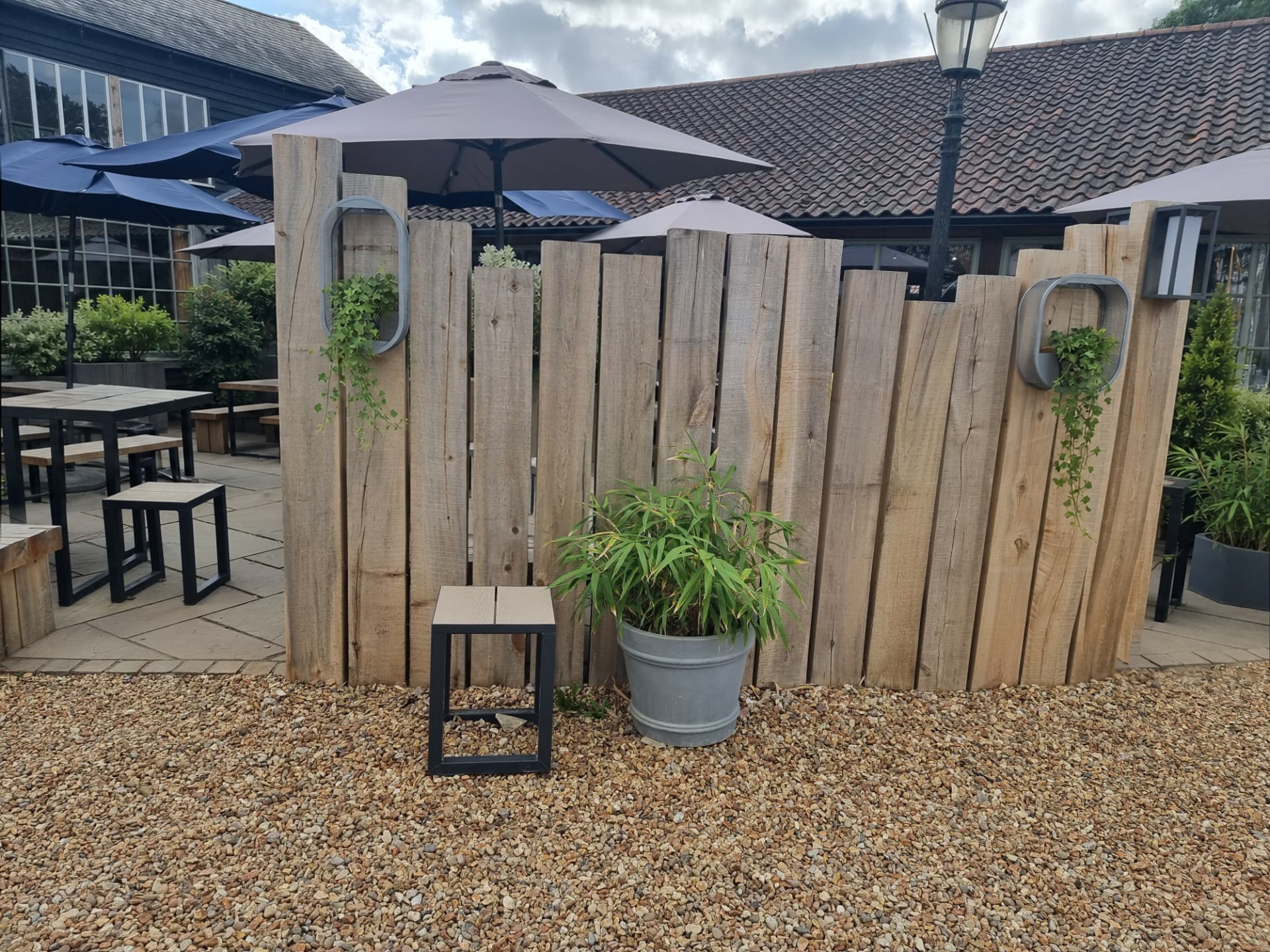 Staggered-height screen of rough-sawn timber sleepers with two wire-mesh wall planters of trailing ivy, screening a paved outdoor seating area