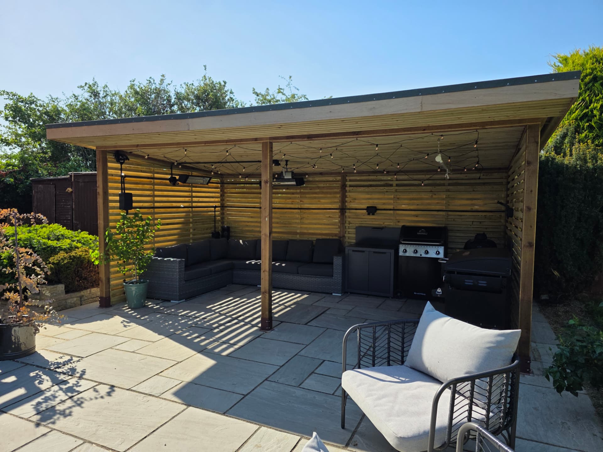 Timber outdoor shelter with a mono-pitch roof, slatted privacy walls and a stone slab floor, fitted with festoon lights and heaters