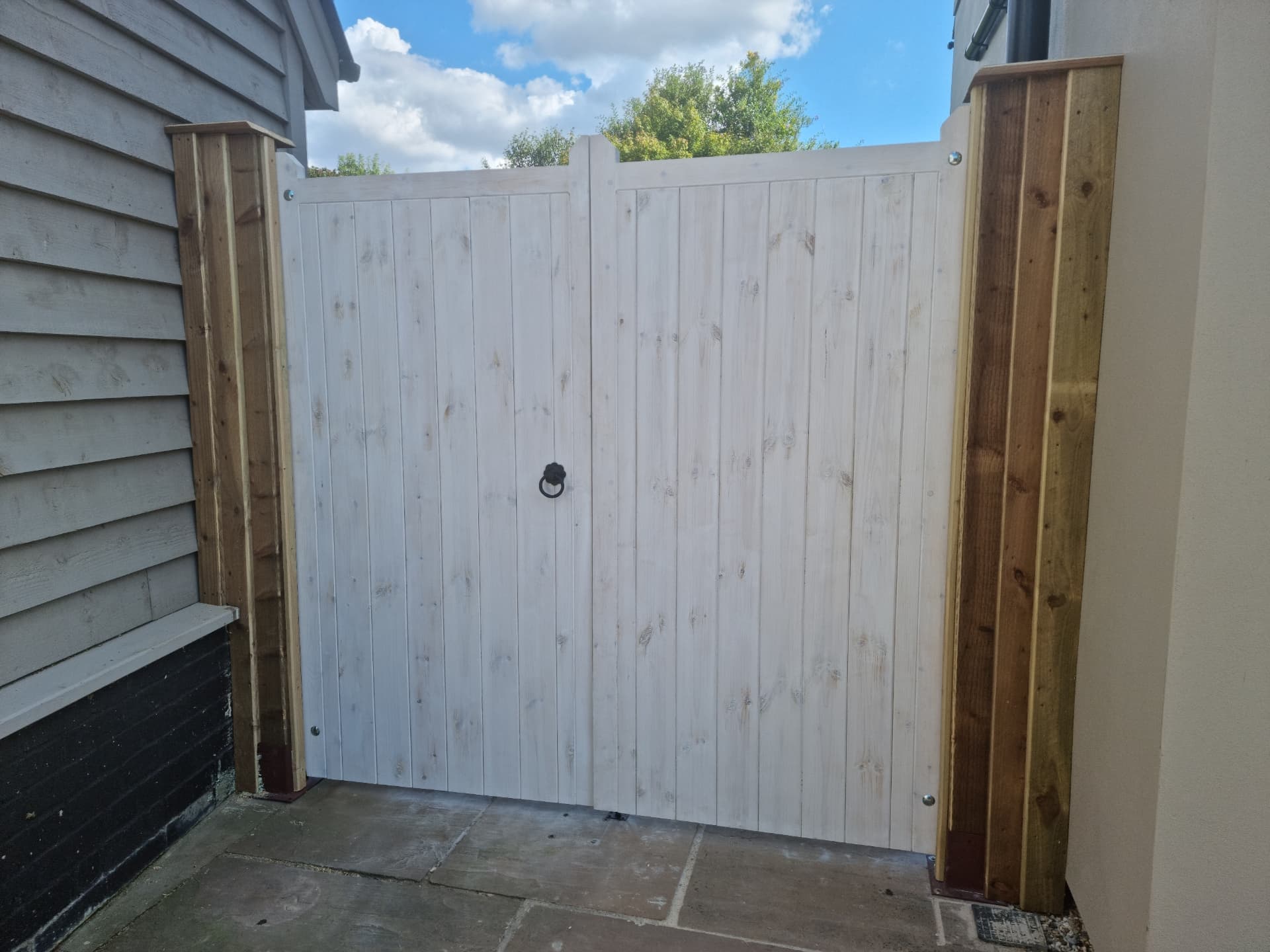 Whitewashed timber double gate with vertical boards and a black ring latch, hung between stained posts at the end of a stone-flagged passage