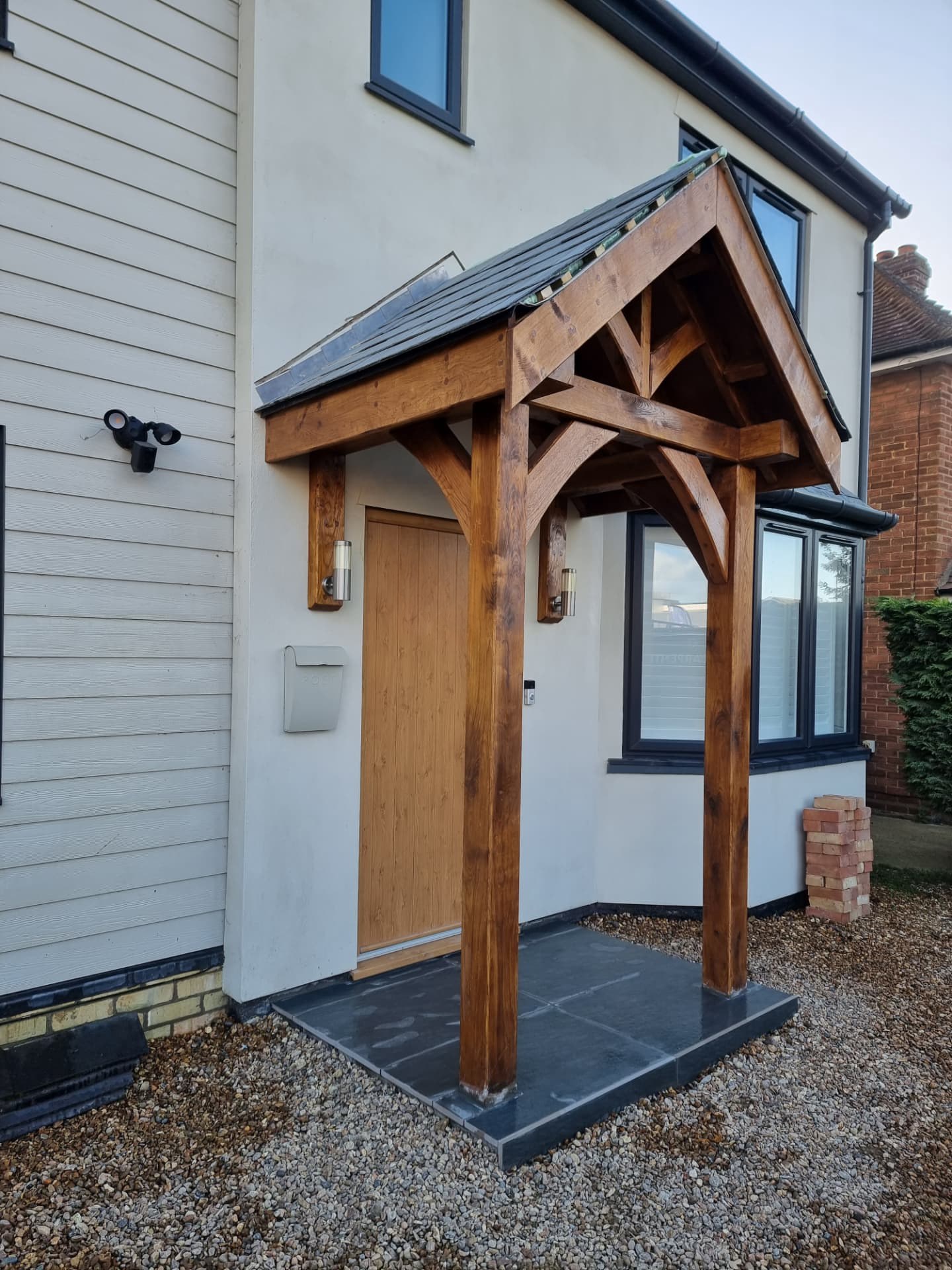 Timber porch with a pitched gable roof, curved braces and slate roof tiles, built against a rendered wall with cream lap siding and a light oak door
