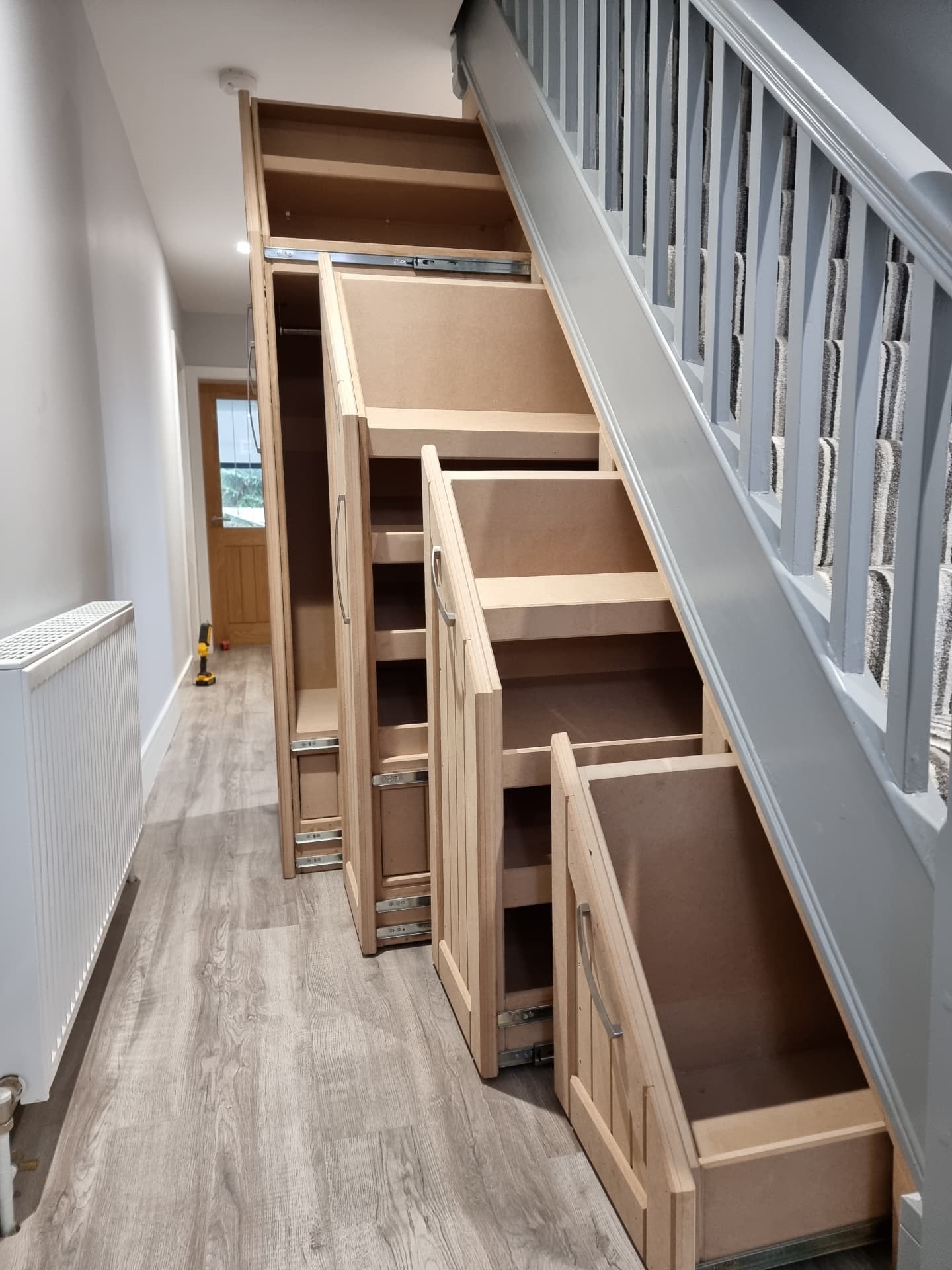 Four graduated pull-out cabinets built under a staircase, from a tall wardrobe section with a hanging rail down to tiered shoe shelves